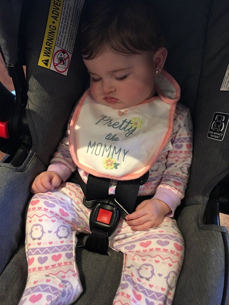 Sleeping child strapped into a car seat, wearing a pink onesie and bib with “Pretty as a Princess, Mommy” text.