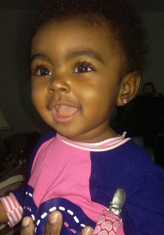 Smiling toddler in a purple and pink sweater, looking up with tongue out against a dark background