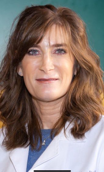 Close-up portrait of a person with wavy brown hair, wearing a white coat, against a teal background