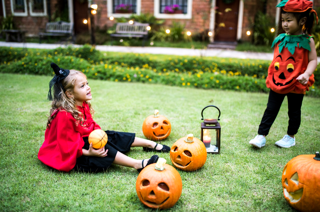 Children in Halloween costumes sit on a lawn with pumpkin decorations and a lantern