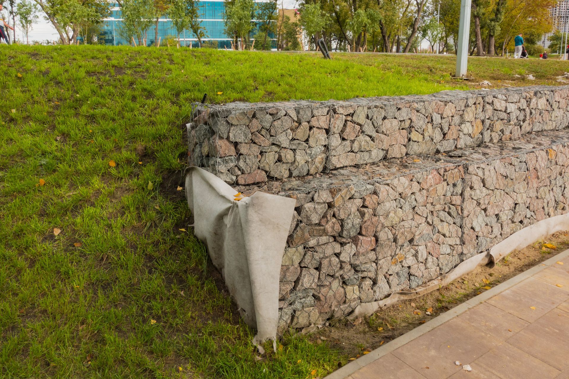 Stone retaining wall with a torn concrete corner beside a paved path and grassy slope