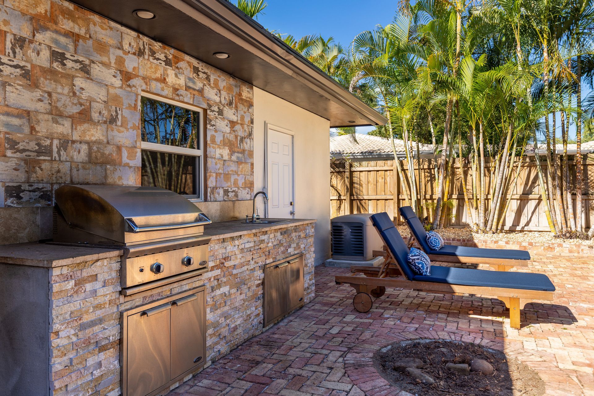 Backyard patio with stone barbecue grill, lounge chairs, and bamboo fencing in sunlight