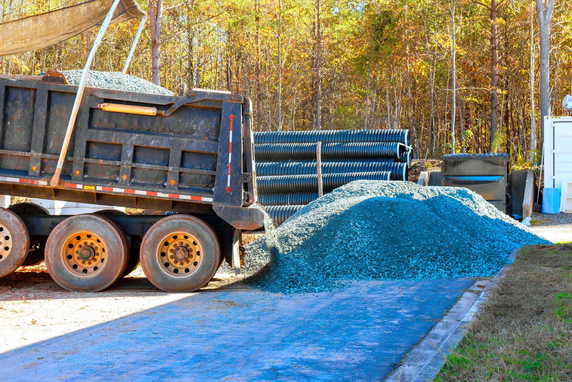 Dump truck unloading blue gravel beside a road in a wooded area