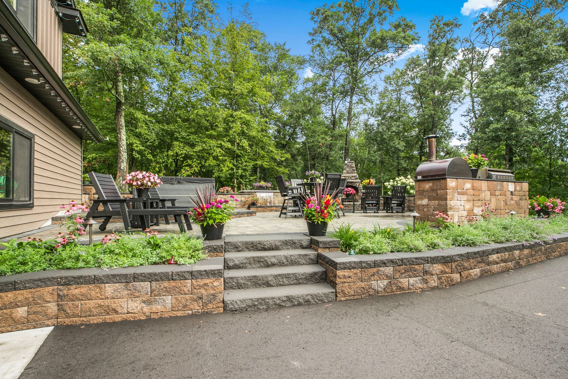 Stone patio with steps, flower beds, and outdoor seating beside a house under trees