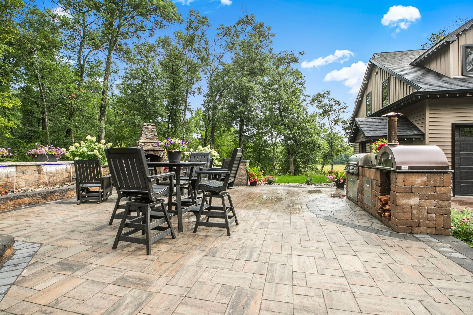 Stone patio with black table and chairs beside a house, bordered by brick planters and trees under a blue sky