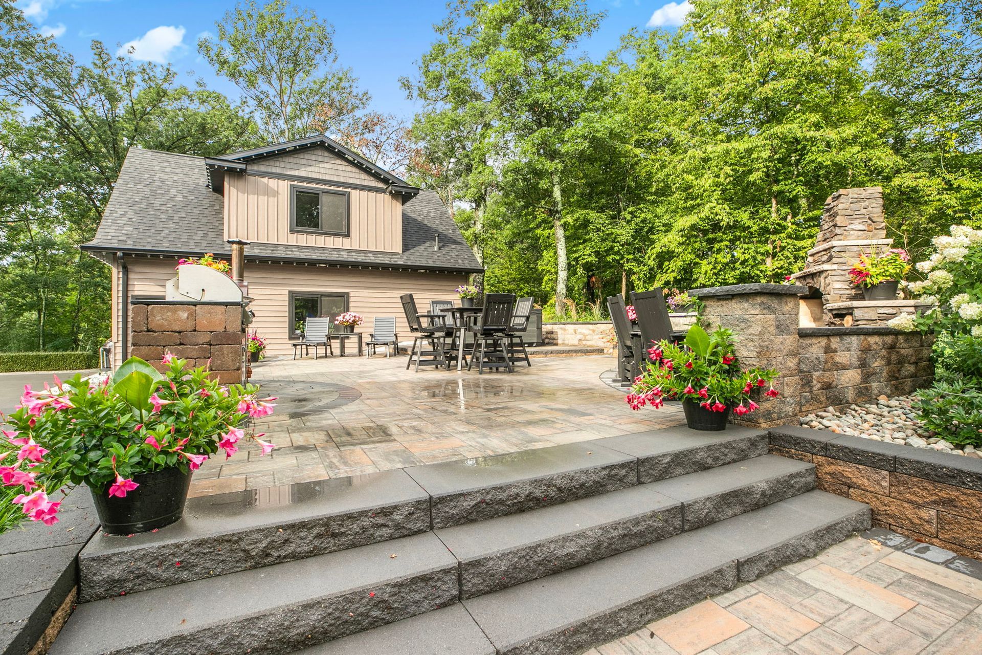 Stone patio with steps, outdoor dining area, pink flowers, and a cottage amid green trees