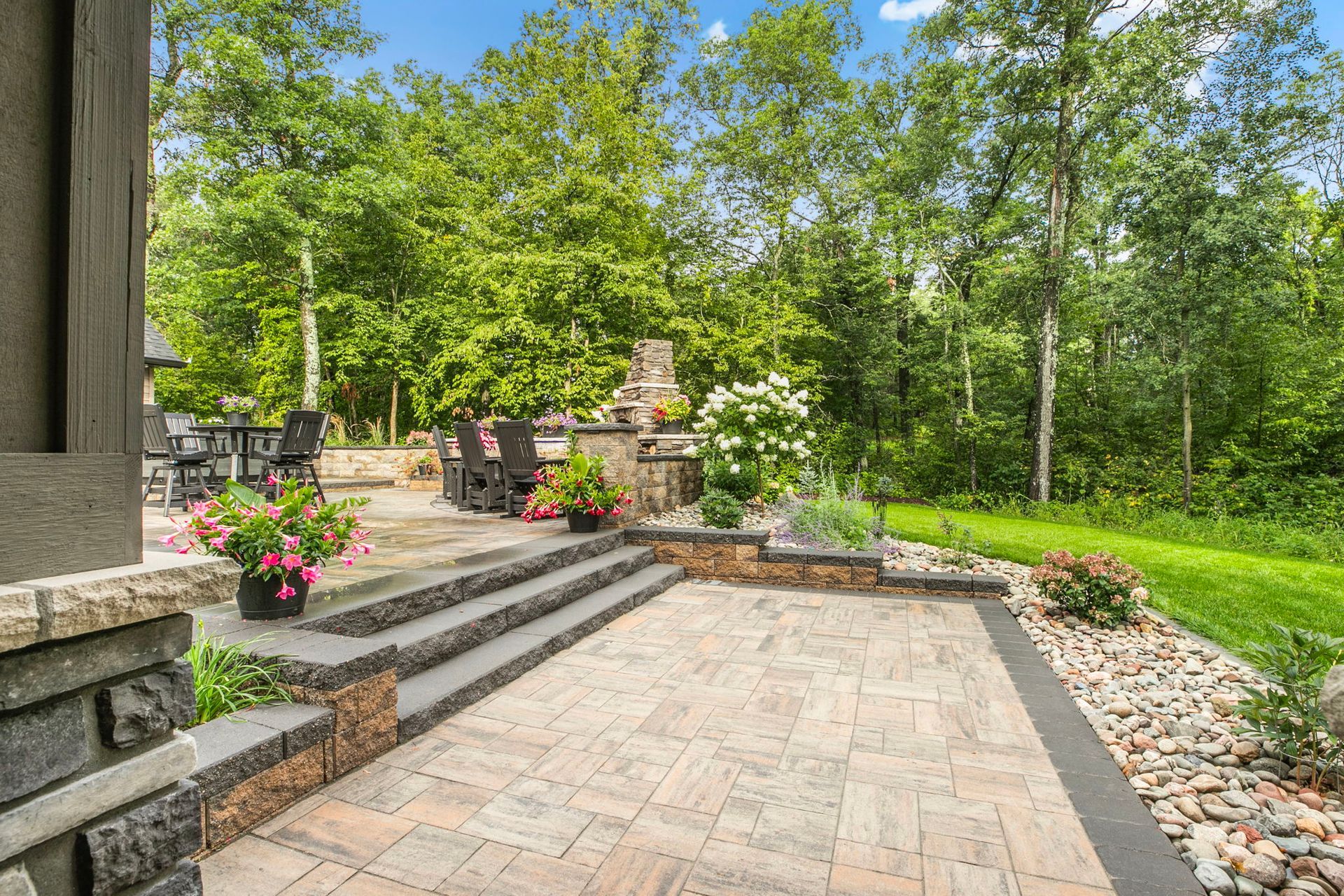 Stone patio and steps in a landscaped backyard with potted flowers, retaining wall, and trees.