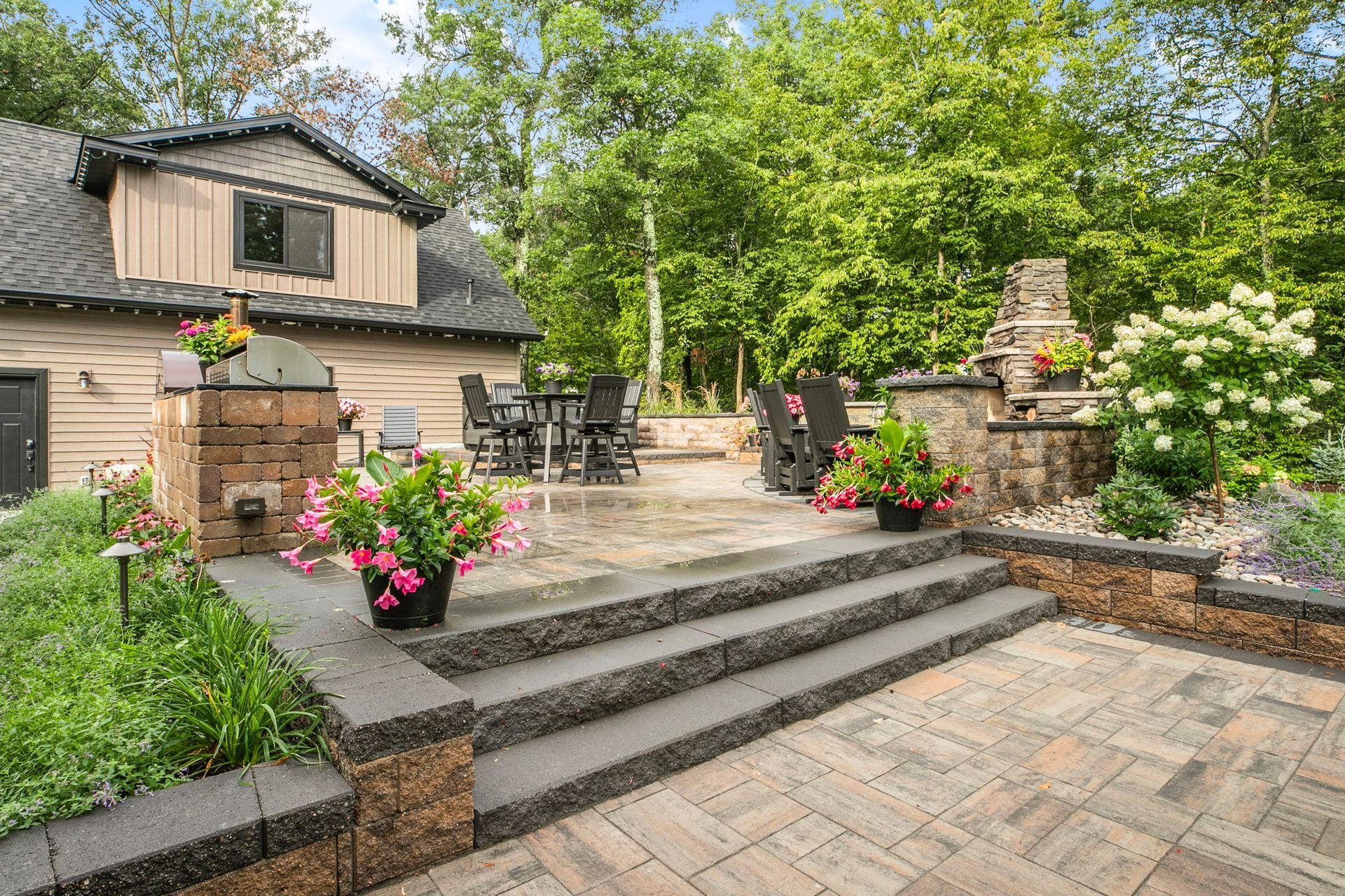 Stone patio steps with potted flowers, outdoor seating, and a brick fireplace in a lush garden.