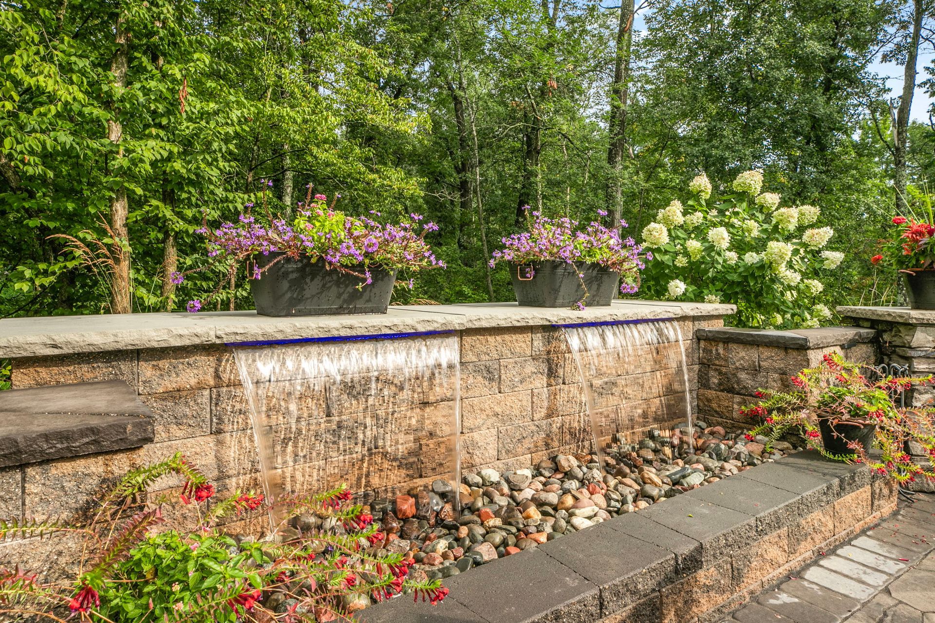 Stone garden waterfall with pink and white flowers and lush green trees.