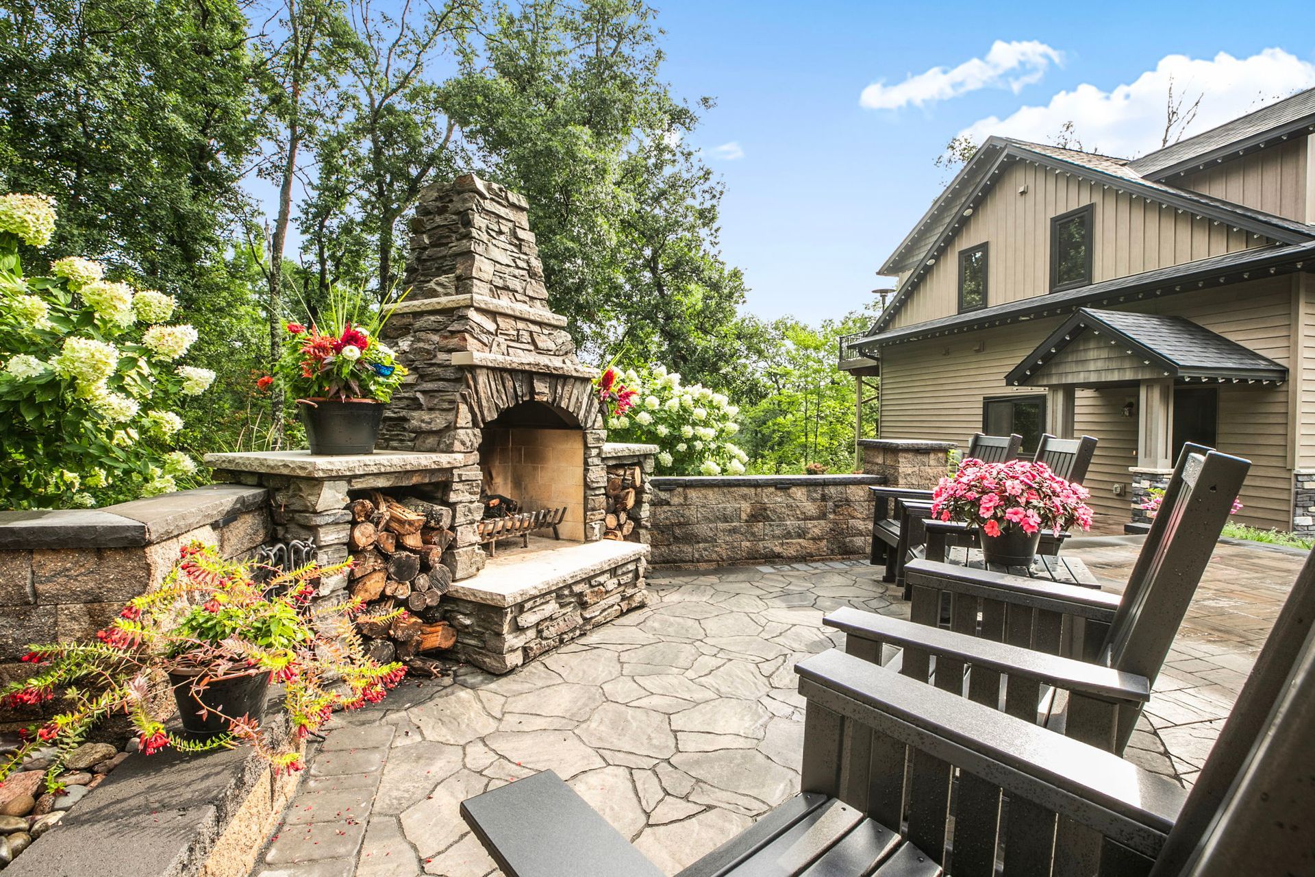 Stone patio with fireplace, flower planters, and Adirondack chairs beside a house