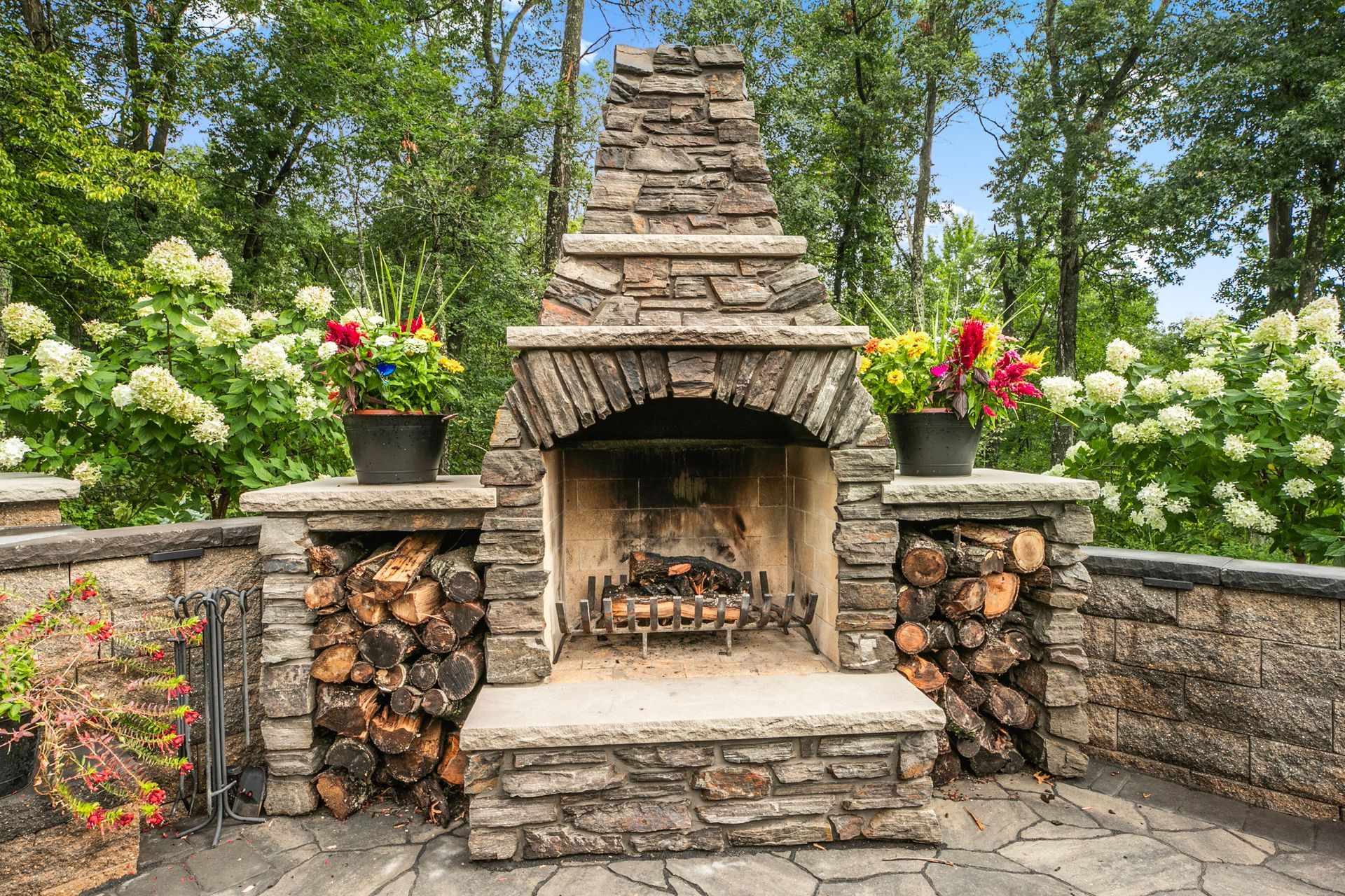 Stone outdoor fireplace with stacked firewood, flower pots, and white hydrangeas in a garden patio