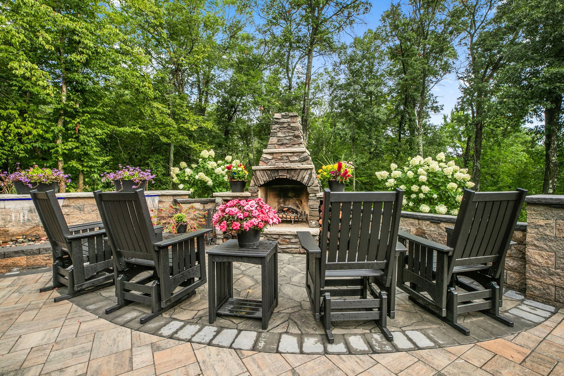 Patio seating area with rocking chairs around a stone fire pit, surrounded by flowers and trees