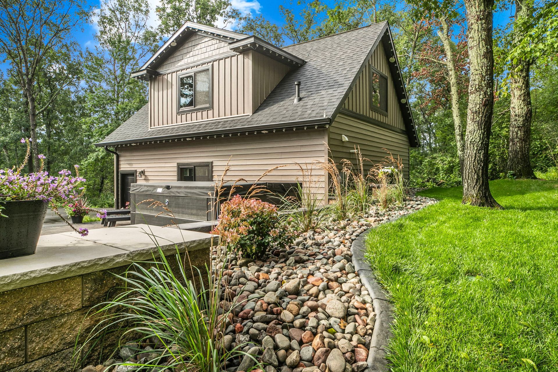 Two-story beige house with a stone-lined garden bed, green lawn, and trees in a sunny yard