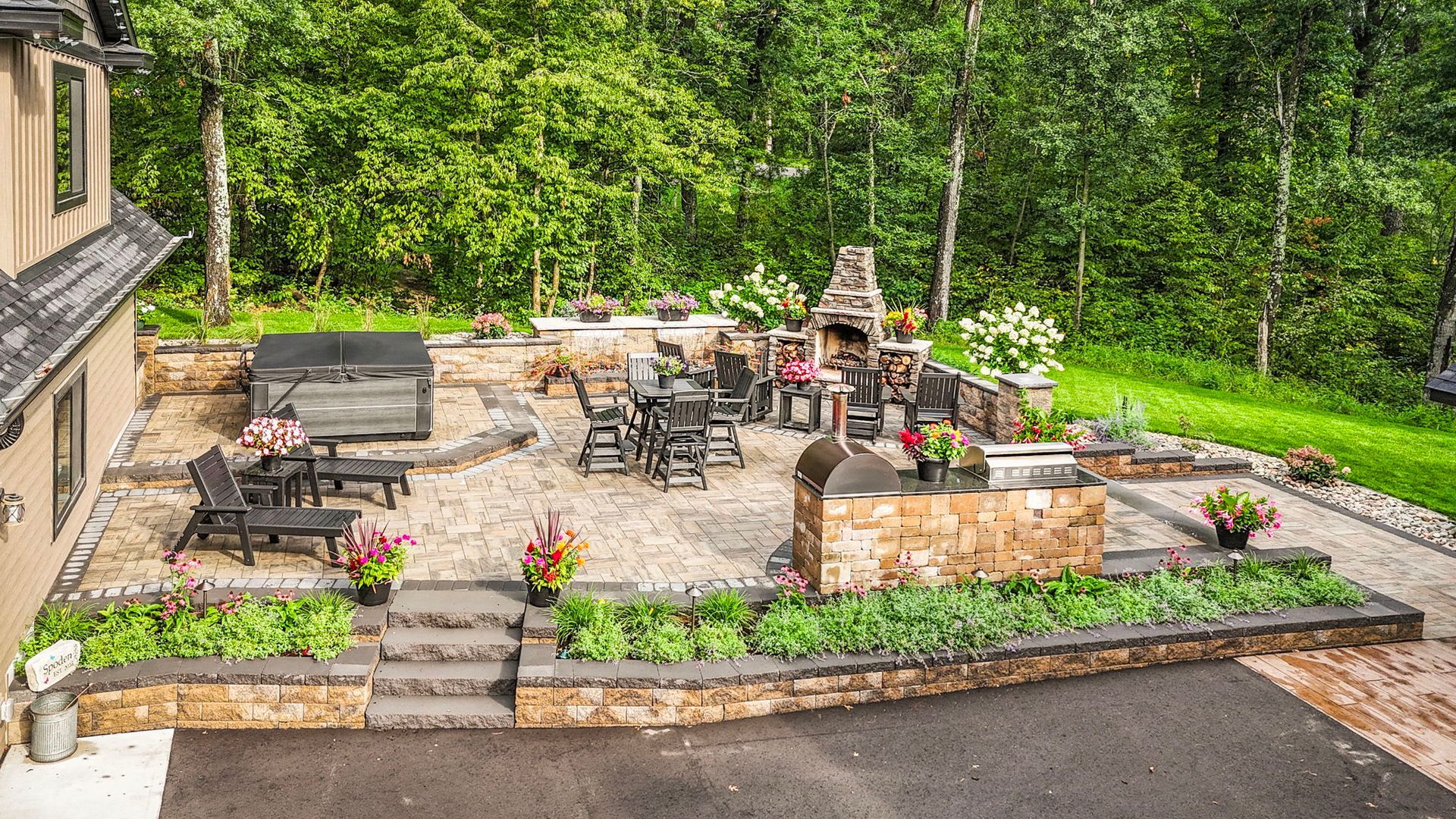 Backyard patio with stone seating, fireplace, potted flowers, and lush green garden backdrop