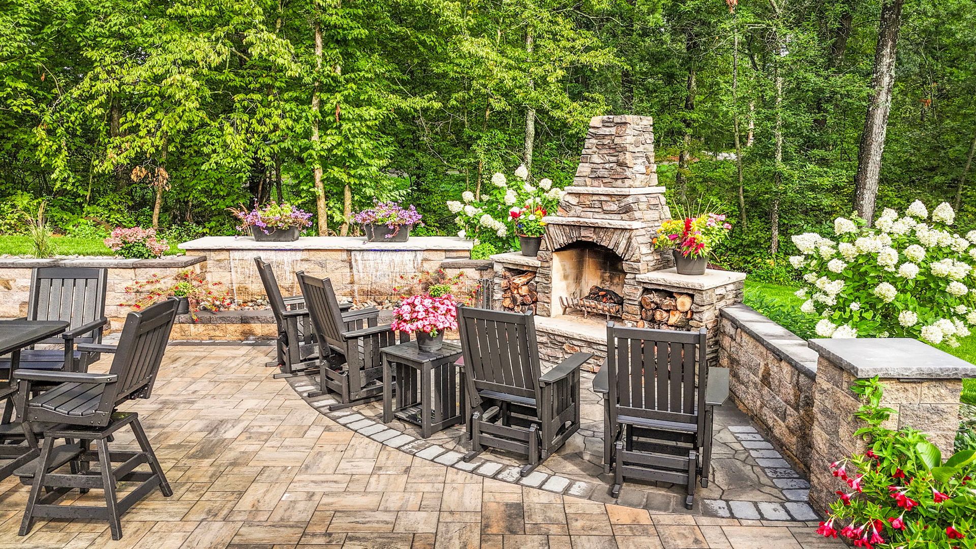 Stone patio with outdoor tables, black chairs, and a brick fireplace surrounded by flowers and trees
