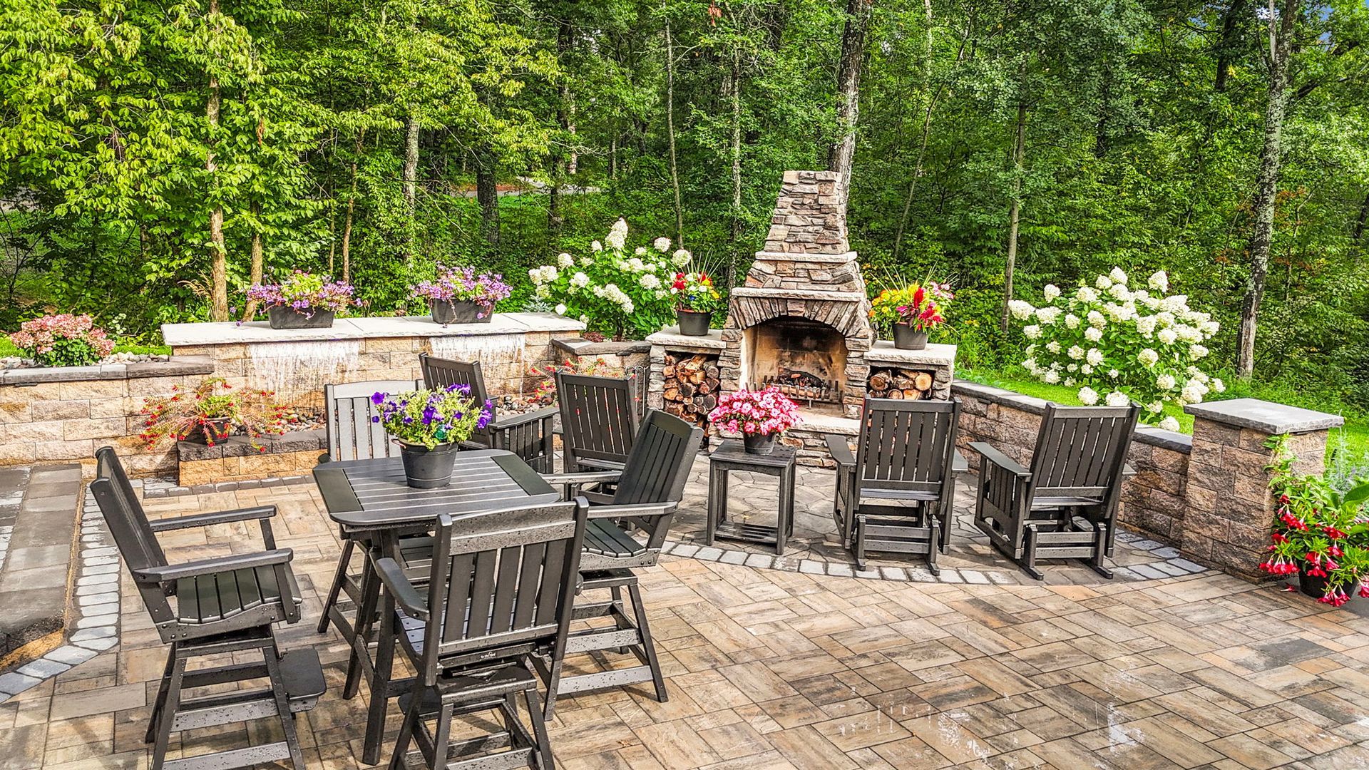 Stone patio with black chairs around a brick fireplace, surrounded by flowers and green trees