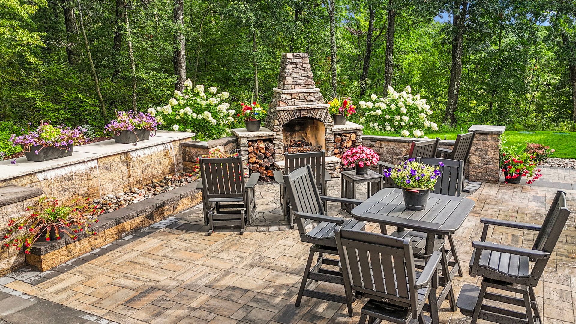 Stone patio with outdoor dining set, brick fireplace, and flower planters in a lush garden