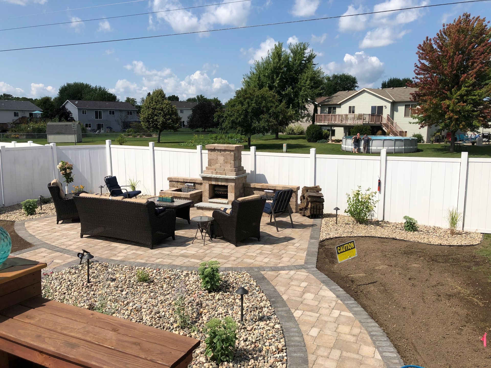 Backyard patio with wicker seating, stone walkway, raised garden beds, and white fence under a blue sky