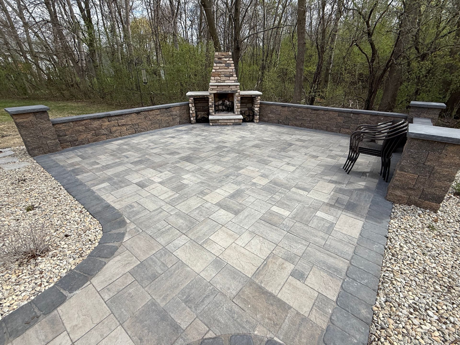 Stone patio with a built-in fireplace, brick walls, and a black chair in a wooded backyard.