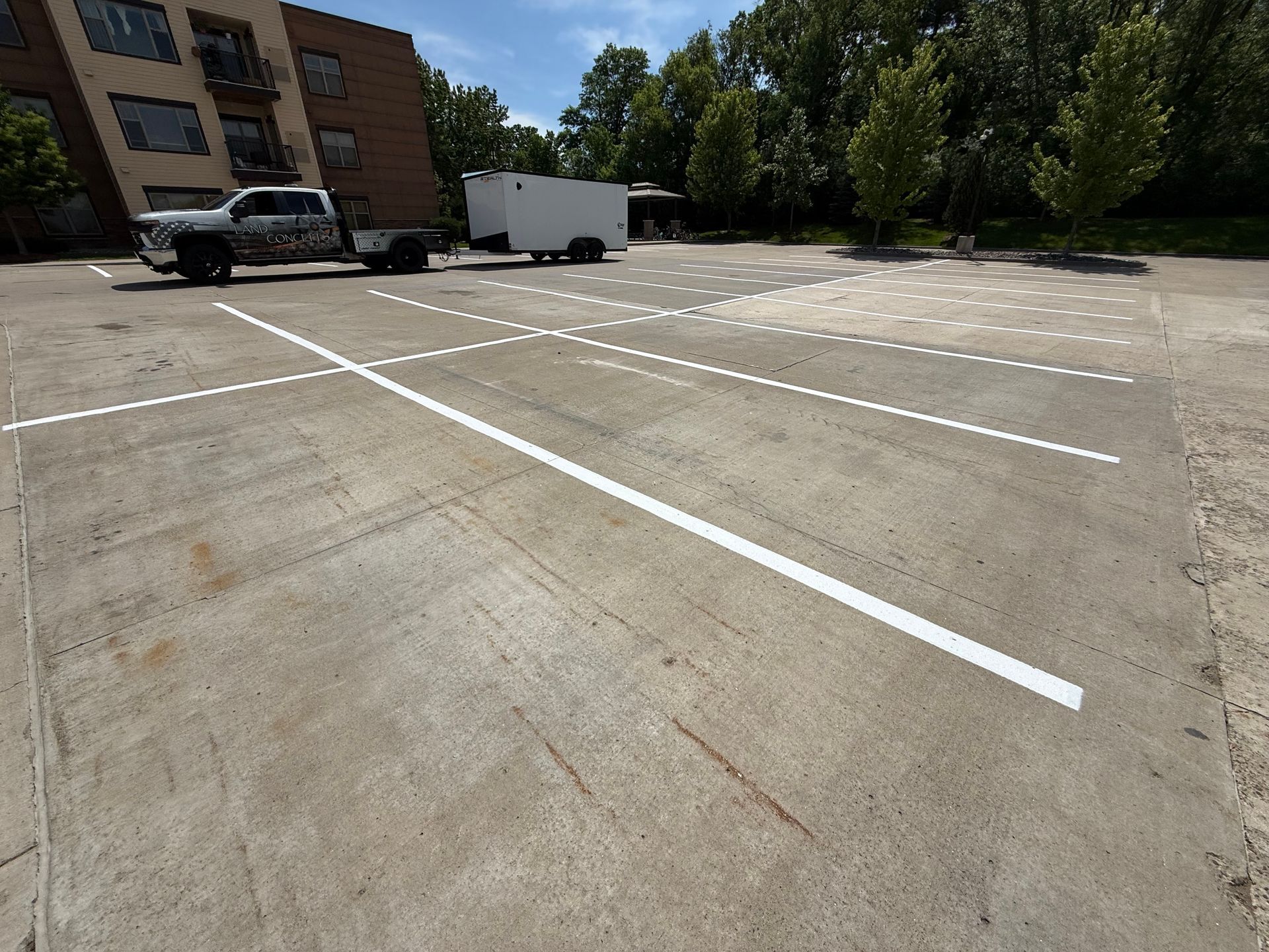 Empty concrete parking lot with white space lines, trees, and a white truck near a building.