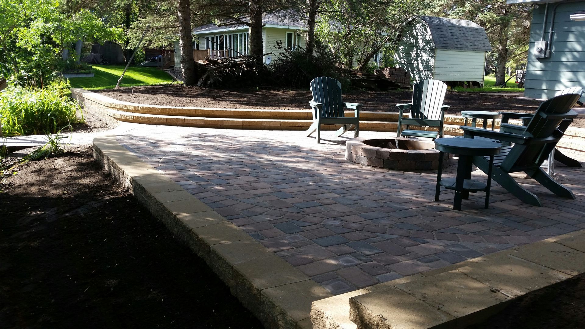 Brick patio with picnic tables and lounge chairs beside a shaded garden and house