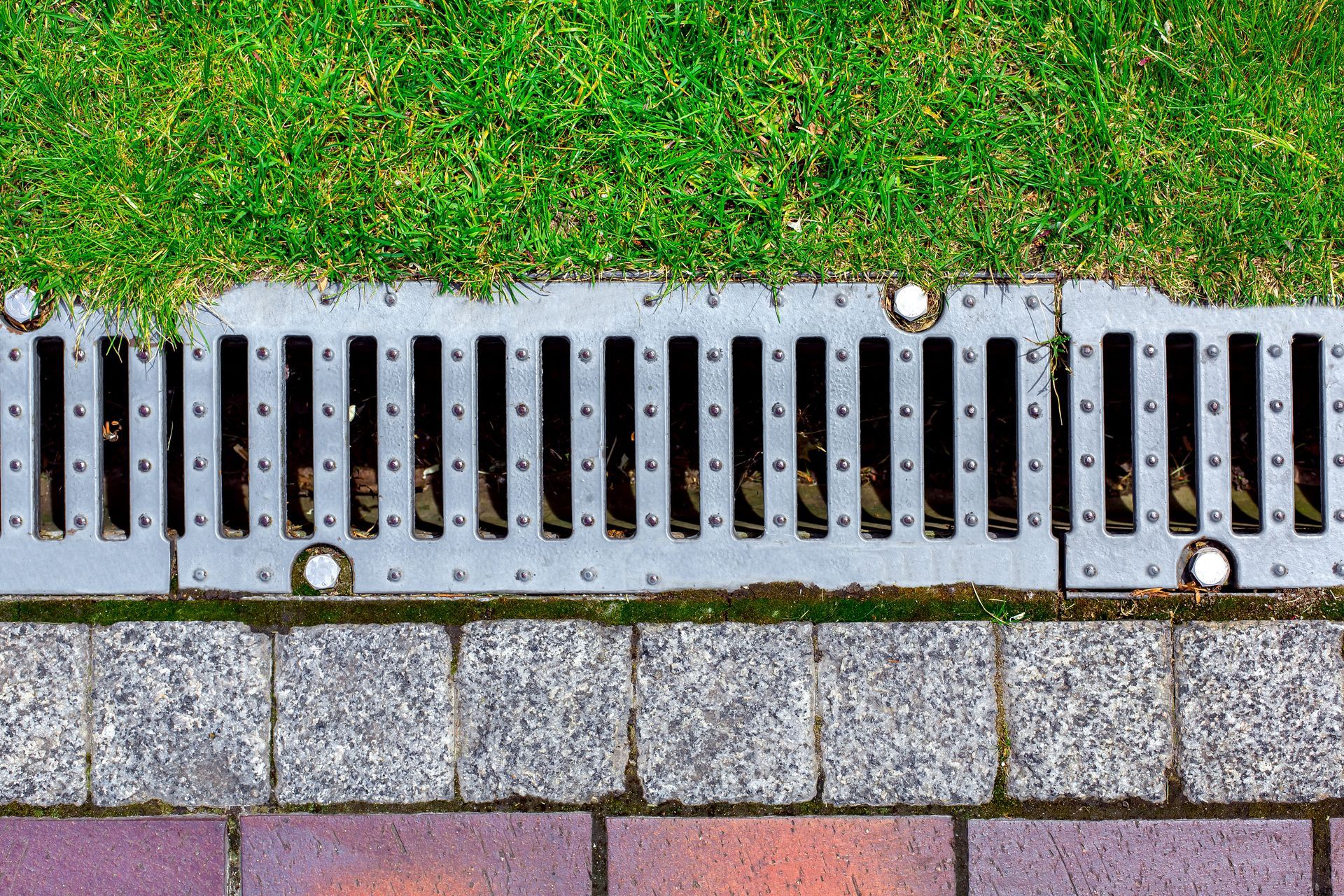 Metal drainage grate beside green grass and stone paving blocks