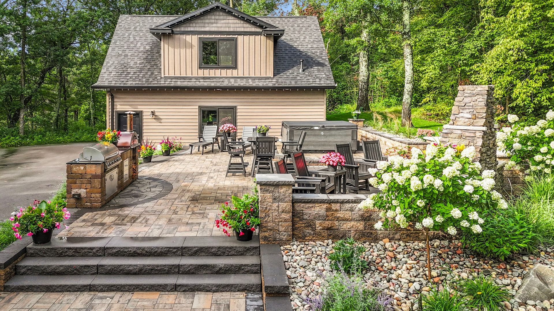 Stone patio with outdoor seating, potted flowers, and a beige cottage-style house surrounded by greenery