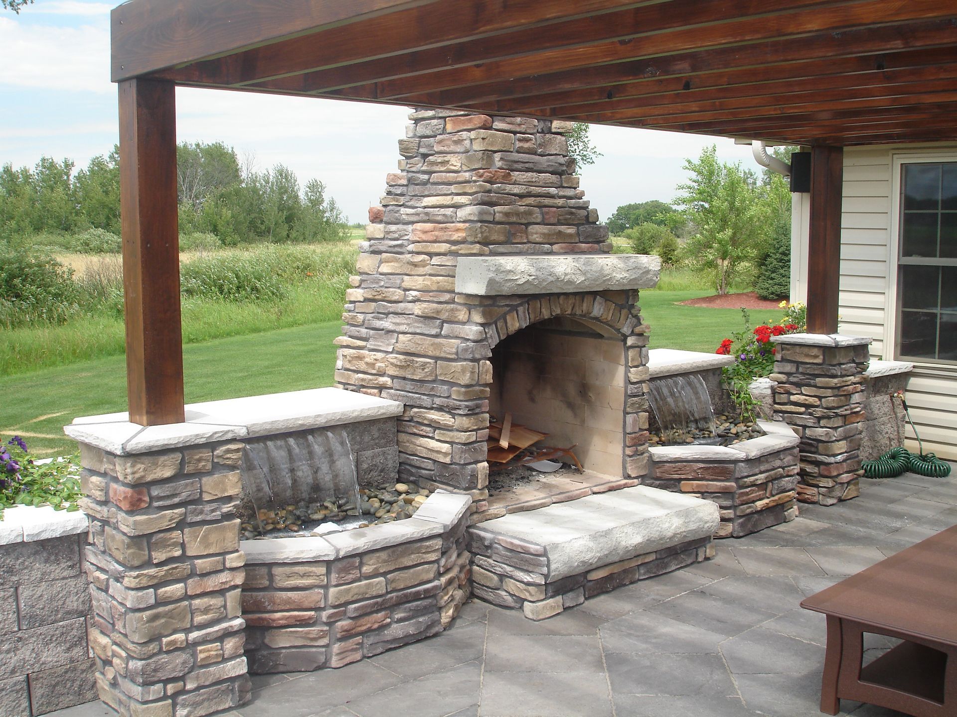 Covered outdoor stone fireplace with built-in benches on a patio overlooking a grassy yard