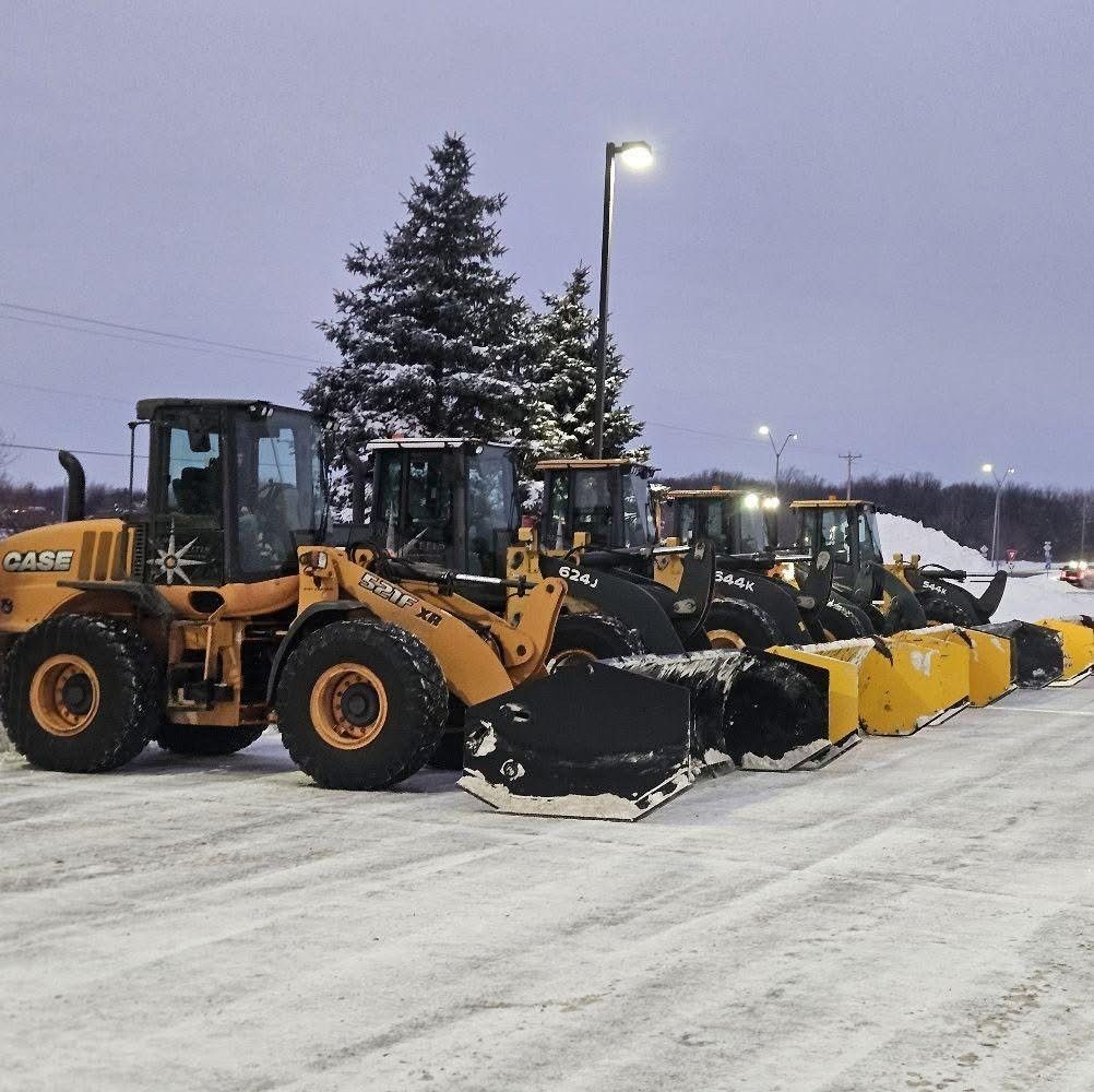 Row of yellow snowplows and loaders lined up on a snowy lot at dusk