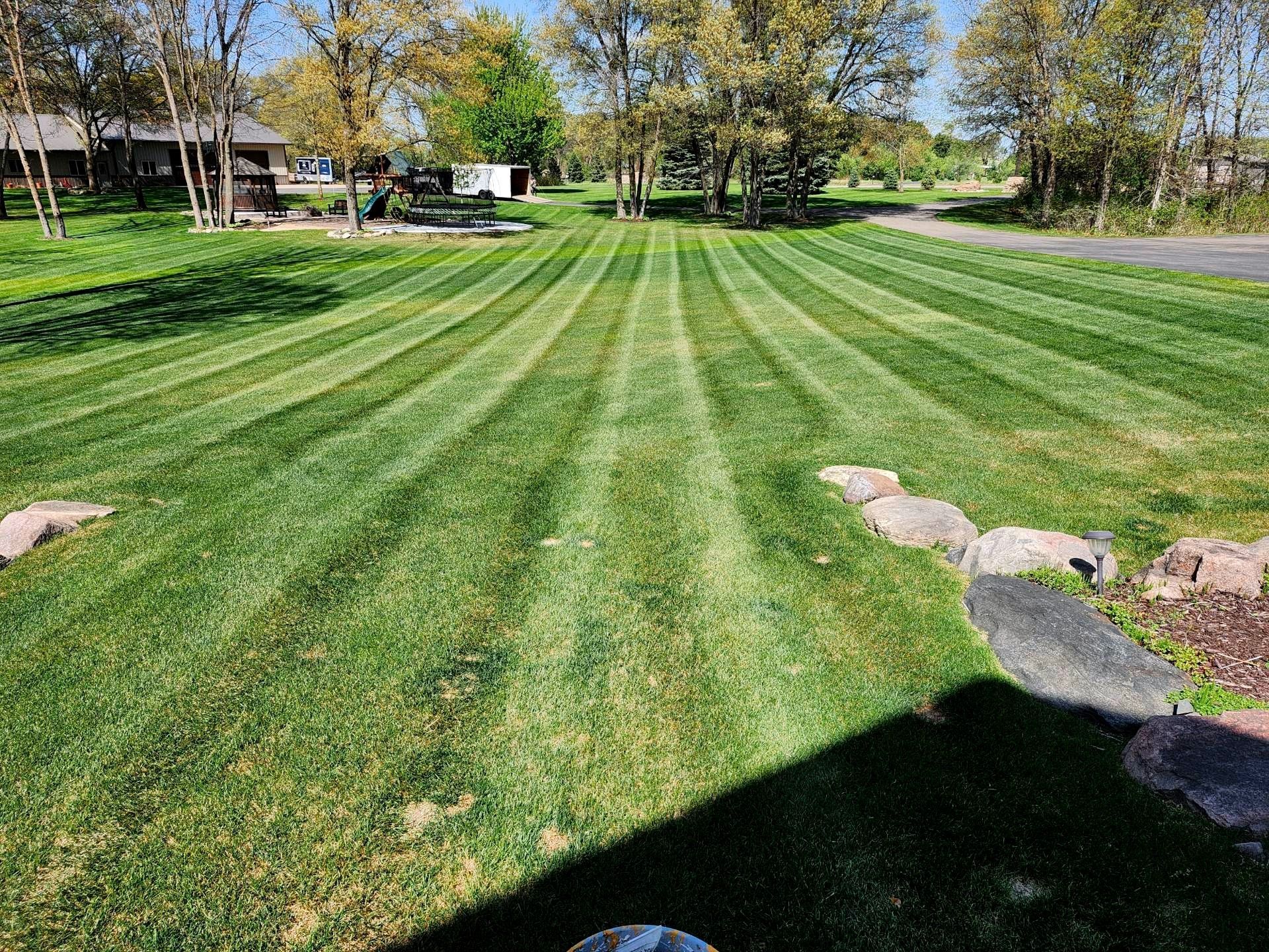 Striped green lawn in a sunny park with trees, rocks, and a house in the background
