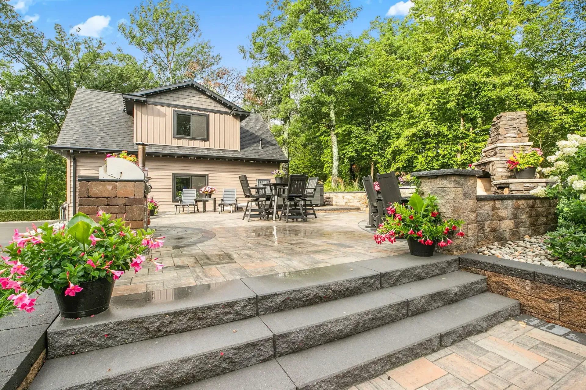 Stone steps leading to a rustic house courtyard with flower pots and lush green trees under a blue sky