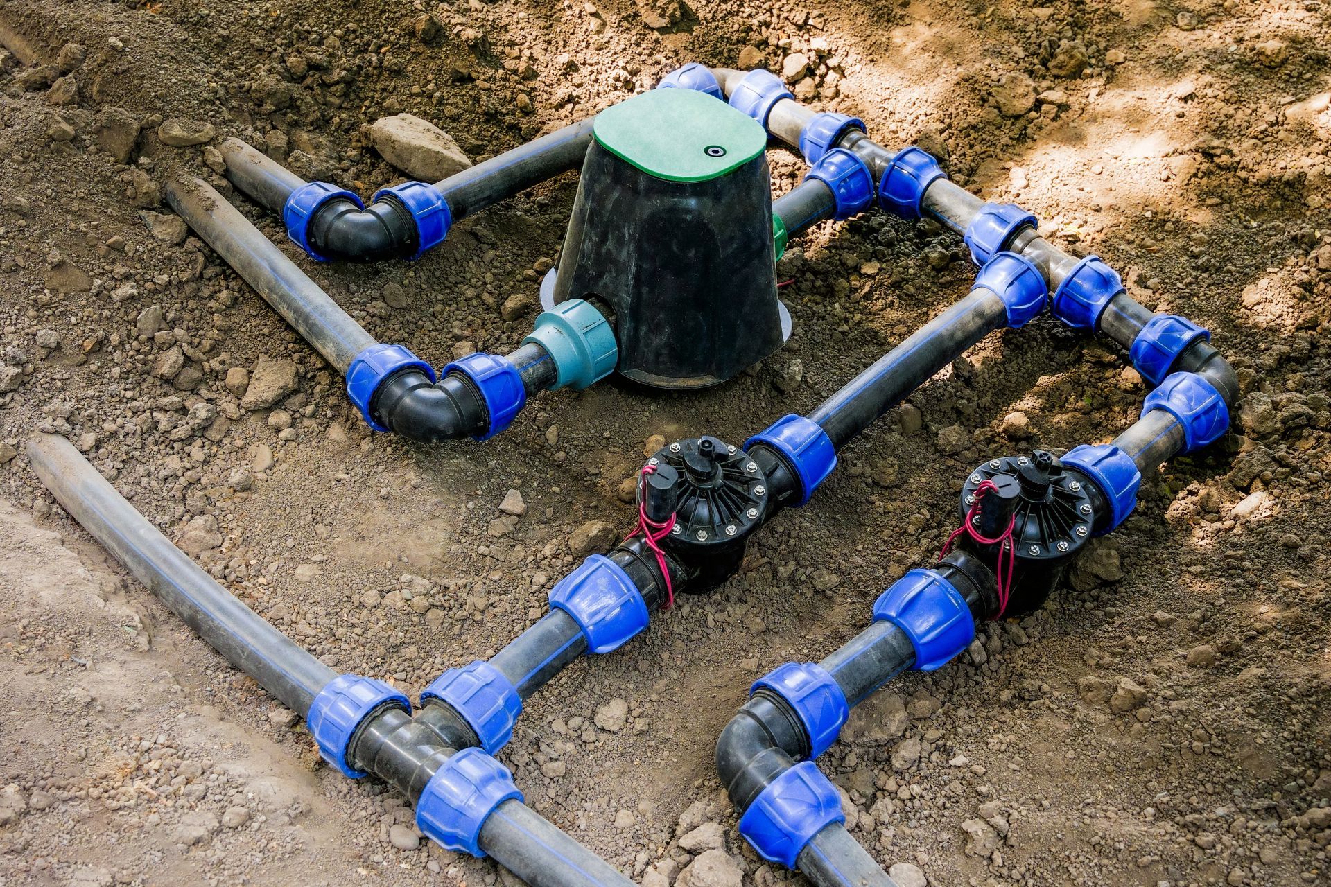 Underground sprinkler plumbing with blue fittings and black pipes around a green valve box in dirt
