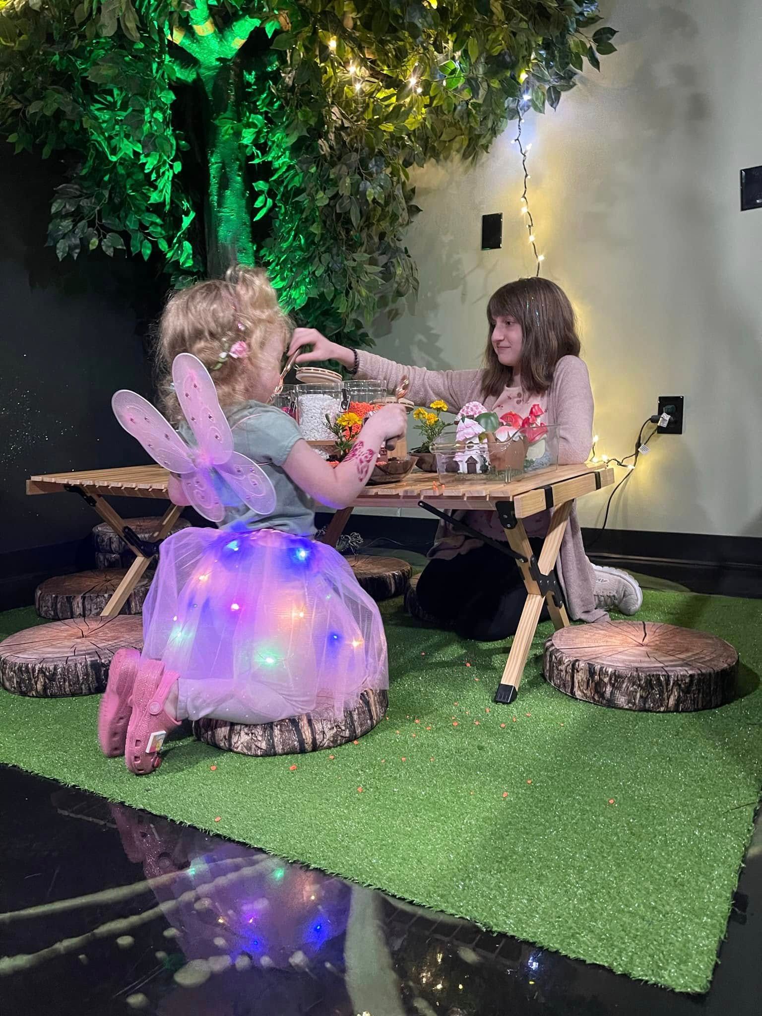 Two little girls are sitting at a table under a tree.