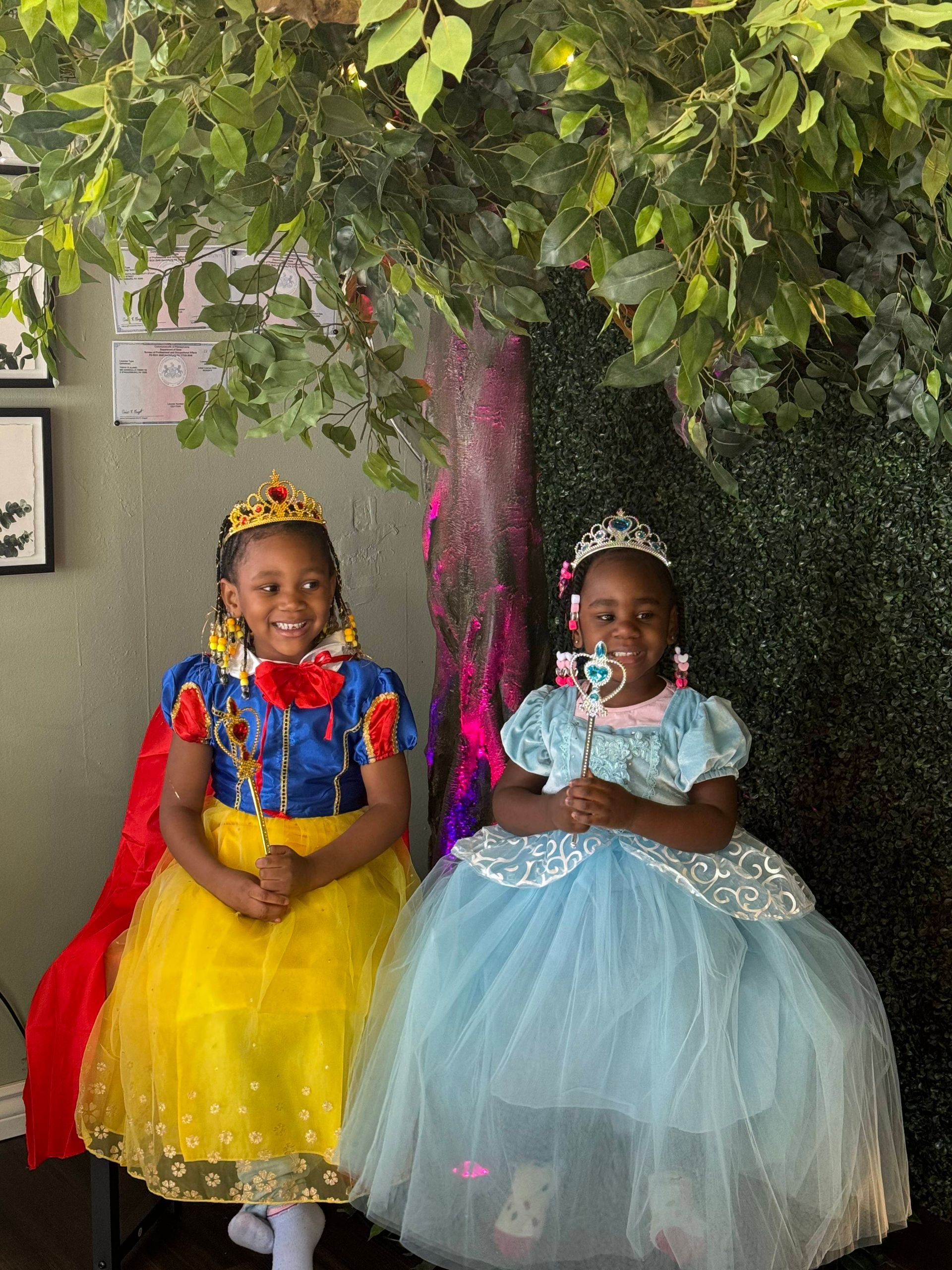 Two little girls dressed as snow white and cinderella are sitting under a tree.