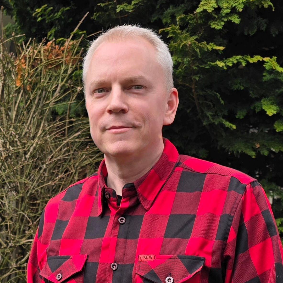 Man with light skin and short gray hair wearing a red and black plaid shirt, smiling outdoors.