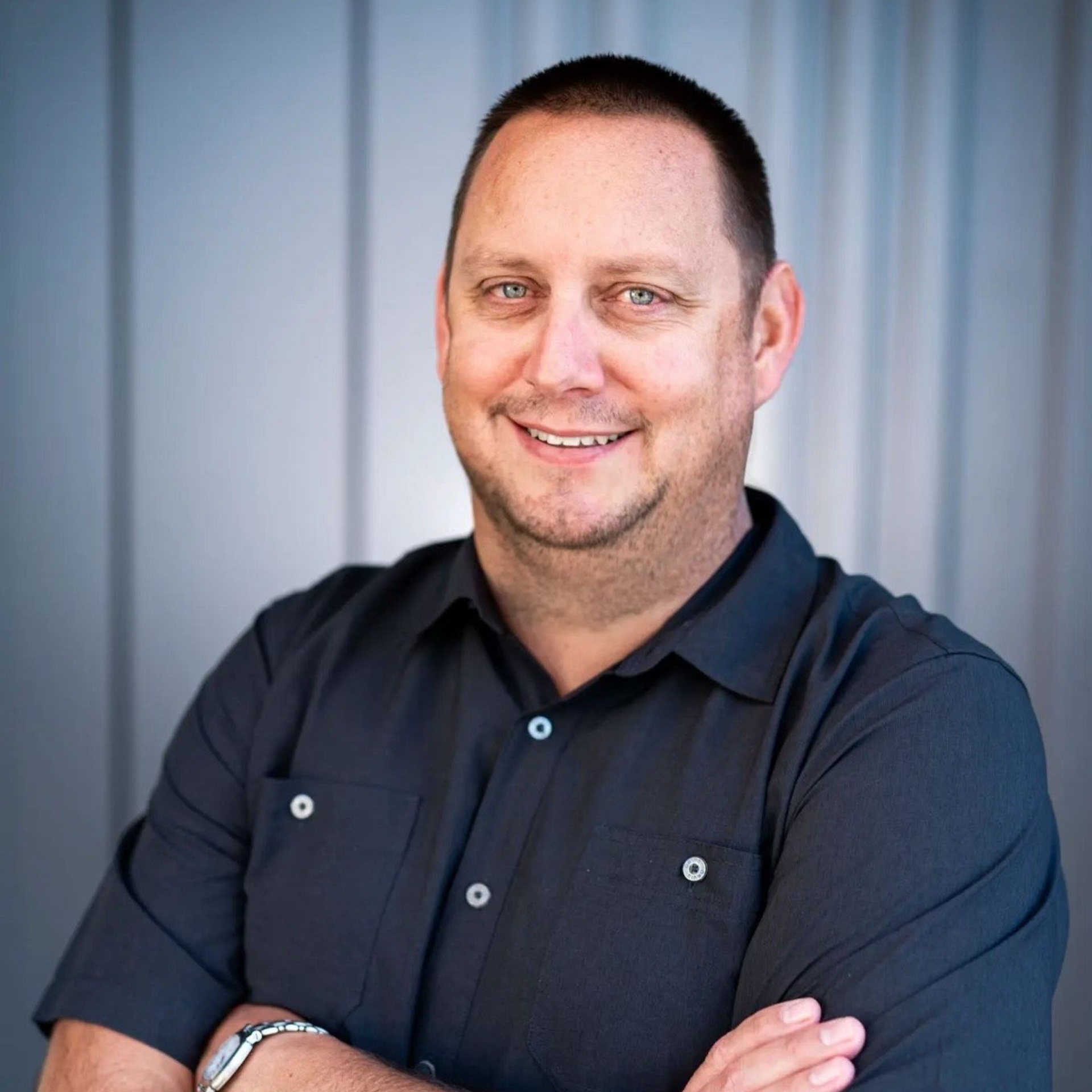 Man in a black button-up shirt with arms crossed, smiling. Background is a gray wall.