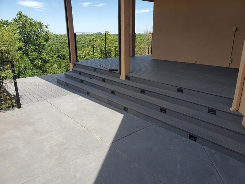 A view of a modern gray deck featuring three wide steps leading up to a covered porch with a wooded background.
