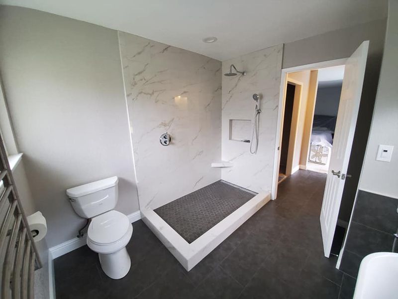 A modern bathroom featuring a walk-in shower with white marble-style walls, a toilet, and an open doorway to a bedroom.