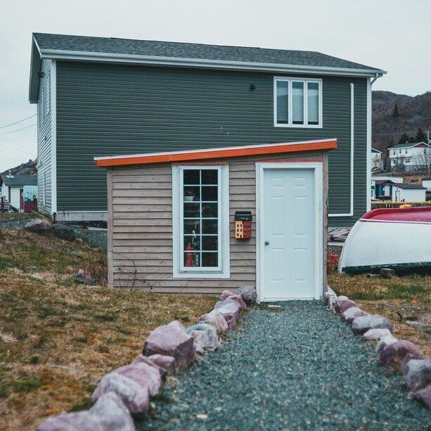 A small, light-wood shed with a white door and orange trim sits in front of a larger, dark grey two-story house.