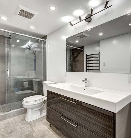 A modern bathroom with a wood-grain vanity, white sink, glass-enclosed shower, toilet, and a large mirror with lighting.