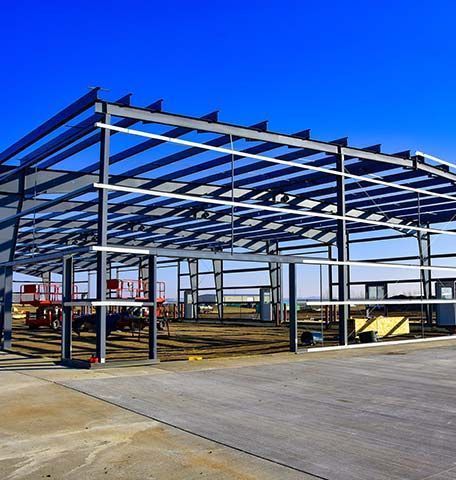 The steel framework of a large, unfinished warehouse stands against a clear blue sky, with construction equipment inside.