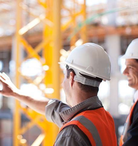 Two construction workers in hard hats and safety vests stand on a job site, with one pointing toward the structure.