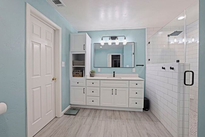 A light blue bathroom featuring a white vanity with a tower cabinet, a mirror, and a glass-enclosed walk-in shower.
