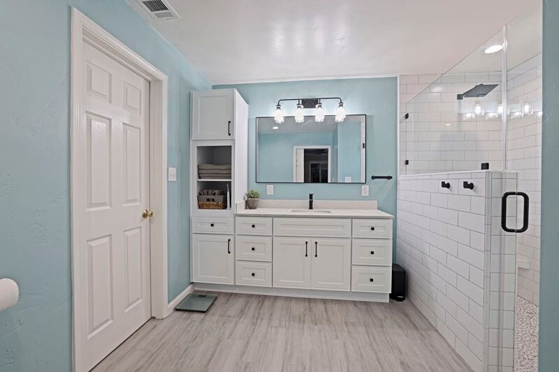 Bathroom with light blue walls, white cabinetry, a single vanity mirror, and a white tiled glass-enclosed shower.