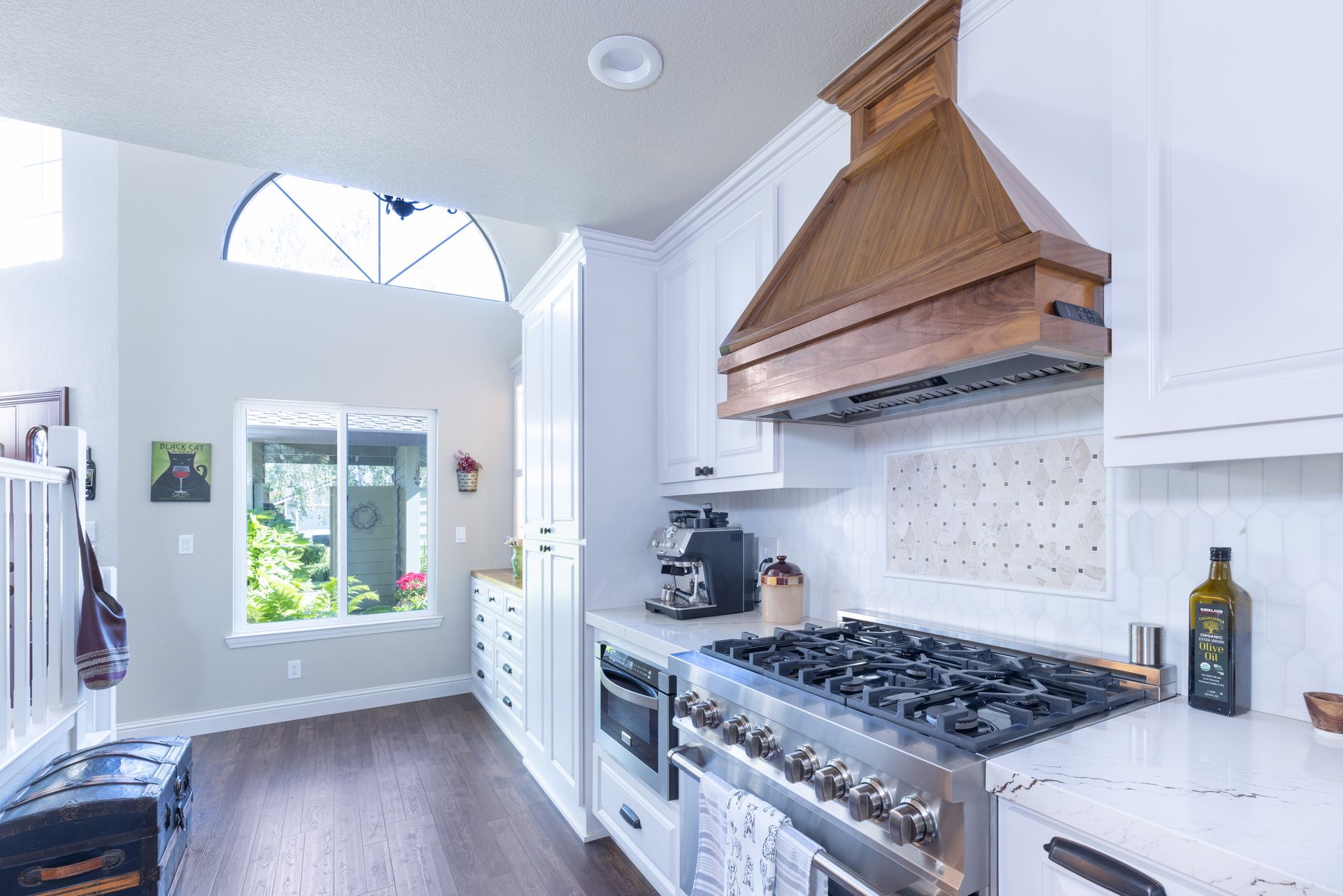 A kitchen featuring white cabinets, a prominent wooden range hood over a gas stove, white counters, and dark wood floors.