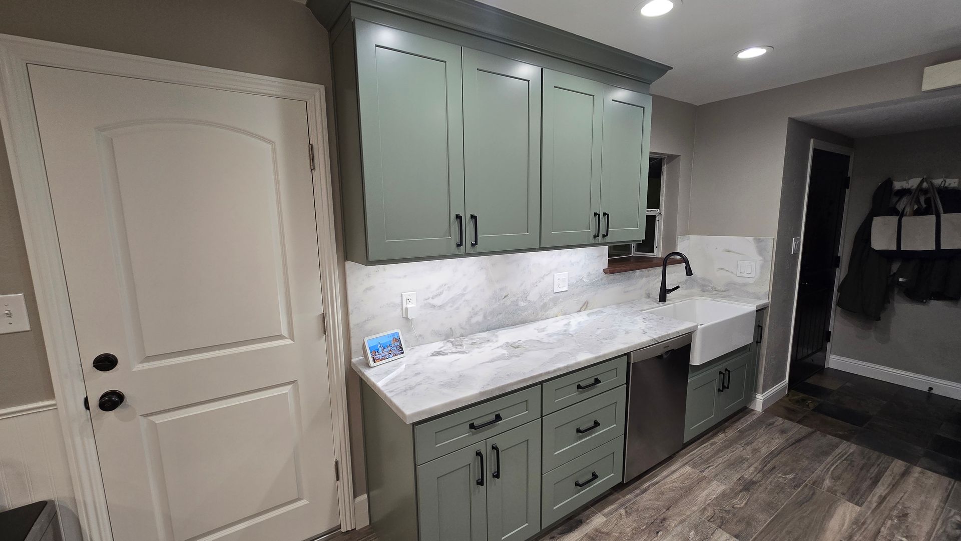 A kitchen area with sage green cabinets, white marble countertops, a white farmhouse sink, and gray plank flooring.