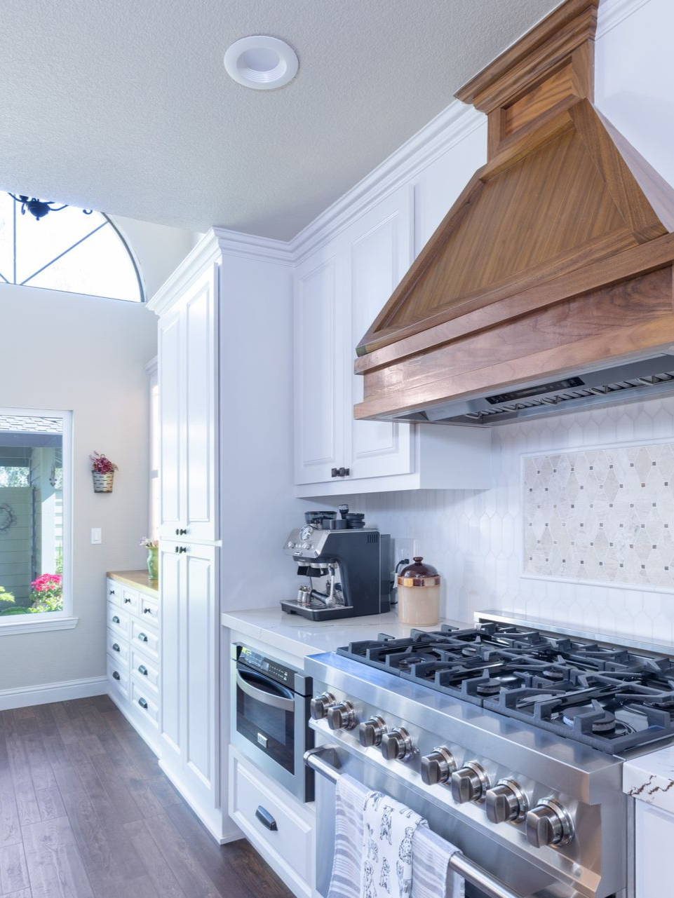 A modern kitchen featuring white cabinets, a prominent wooden vent hood, a stainless steel gas range, and wood flooring.