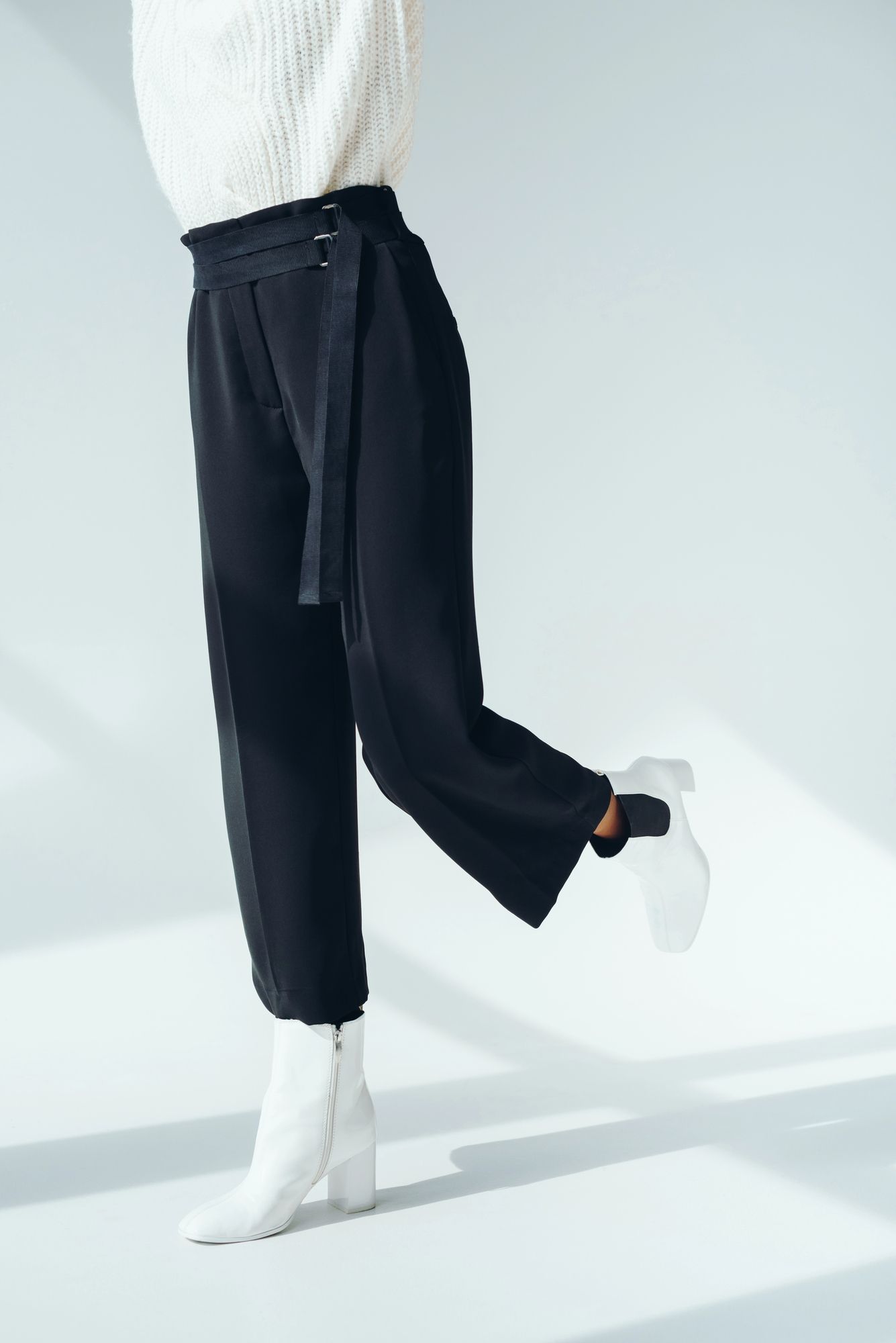 Una persona aparece de pie sobre un fondo blanco, vistiendo pantalones negros de talle alto con cinturón y botas blancas.
