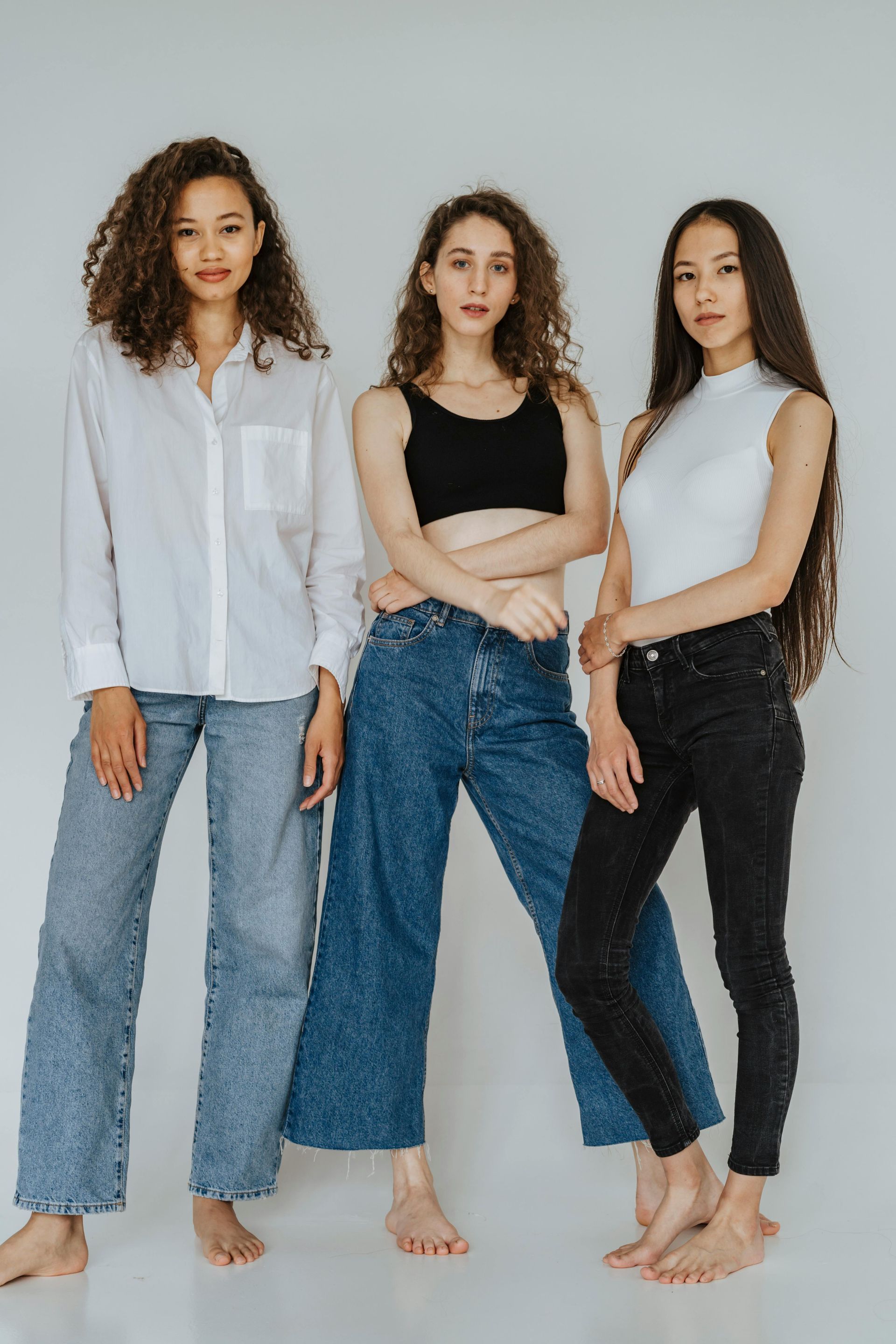 Tres mujeres descalzas, vestidas con vaqueros y camisetas informales, de pie sobre un fondo gris claro.