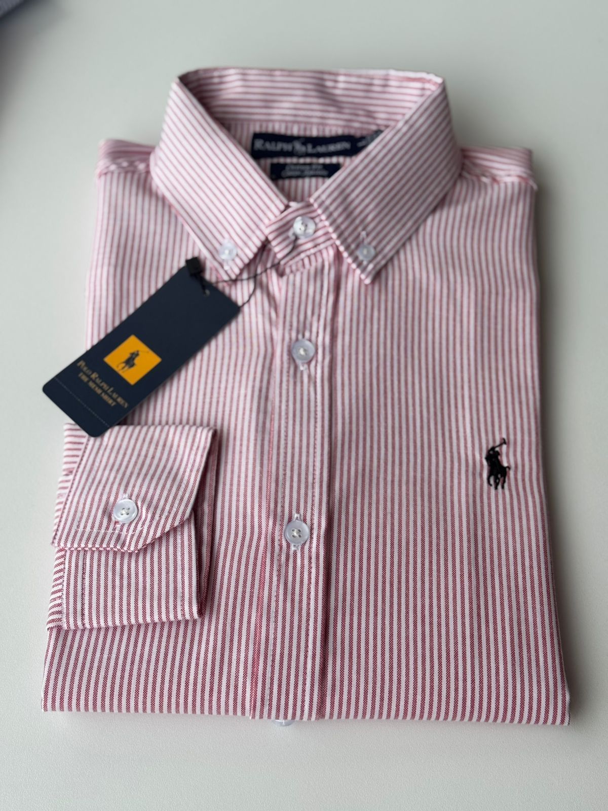 Camisa abotonada de rayas rosas y blancas doblada, con etiqueta y logotipo bordado sobre fondo claro.